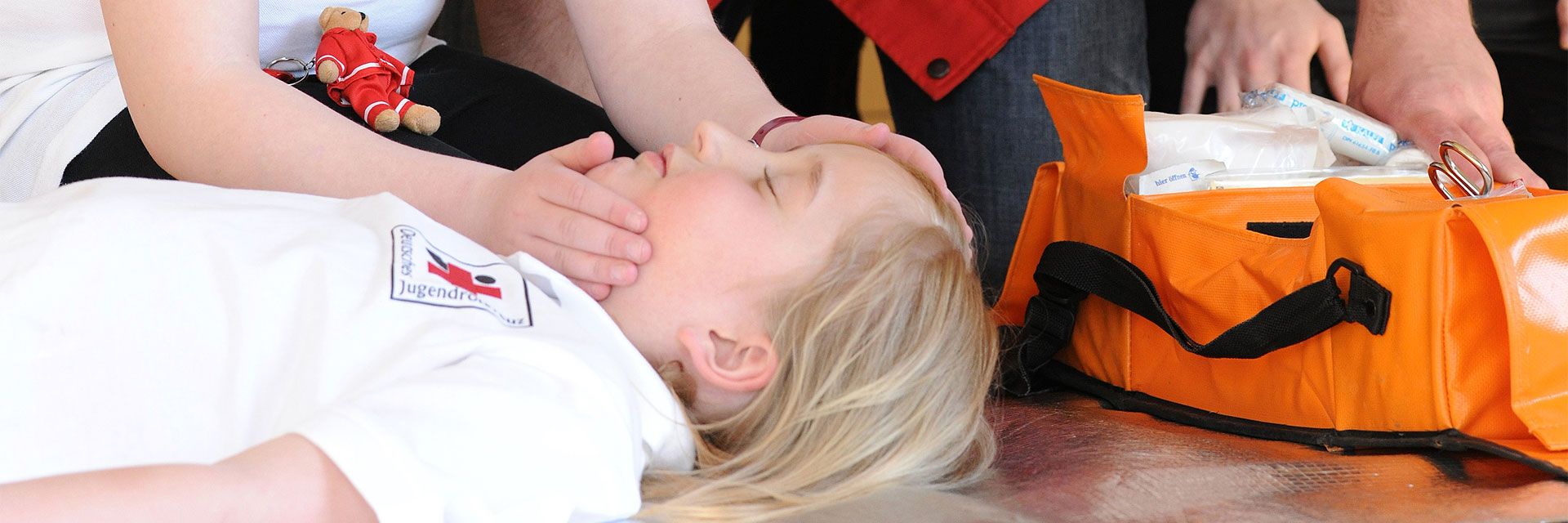 Foto: Bei einer Erste-Hilfe-Übung des Jugendrotkreuzes liegt ein Kind bewusstlos am Boden. Ein anderes Kind überprüft gerade die Atmung. Am Rande des Fotos ist eine Tasche mit Verbandsmaterial zu sehen.